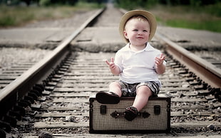 photo of a child sitting on briefcase in the middle of train rail