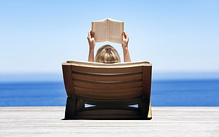 person laying on brown wooden chair reading a book in beach