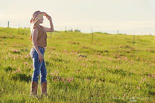 woman wearing brown tank top, cowboy hat, and blue jeans standing in green grasses facing the right side