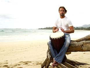 man playing percussion instrument sitting on the rock near the shore