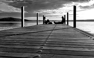 grayscale photo of man and woman sitting on the edge of dock
