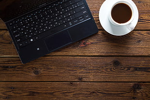 black laptop computer beside white ceramic mug filled with brown liquid on white ceramic saucer