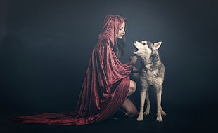 woman in red hood sits in front of gray and white wolf
