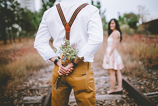 man in white dress shirt hiding flowers in his back near woman in white sleeveless dress