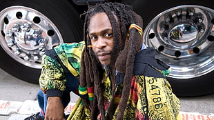 man with dreadlocks wearing multi-colored shirt sitting near chrome vehicle wheel with tires