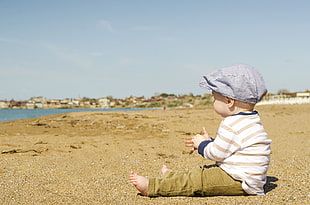 baby in white long sleeve sitting on sand beside beach during daytime