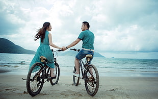 woman in blue sleeveless dress and man in blue shirt sitting on bicycles holding hands near body of water