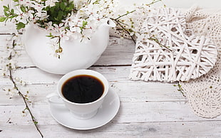 white ceramic tea cup and saucer with black coffee on wooden table near ceramic vase