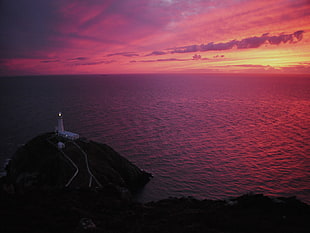 lightened lighthouse on black island near body of water under orange sunset HD wallpaper