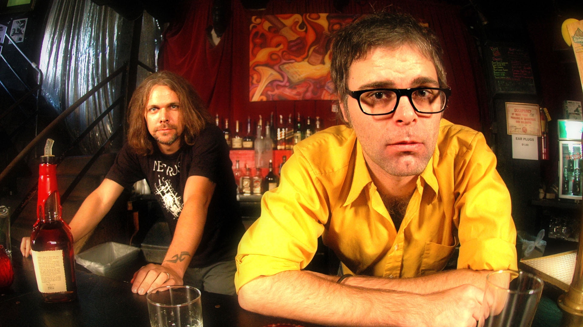 two men leaning on table with glass bottle and drinking glass on top