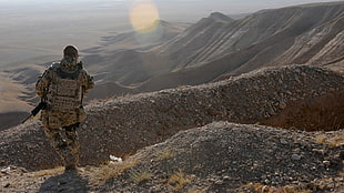 man in military suit carrying black assault rifle standing alone on mountain at daytime