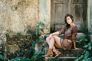 woman in brown long-sleeved dress and pair of brown boots sitting on black concrete stairs near a gray wooden door
