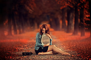 woman in blue denim jacket and black mini skirt holding oval mirror sitting in the middle of pathway in forest