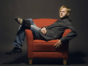 man in black suit jacket with blue jeans sitting on orange fabric armchair