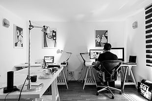 grayscale photo of man sitting on rolling chair in front of computer monitor inside the room