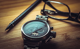 shallow photo of black chronograph watch near black pen and reading glasses
