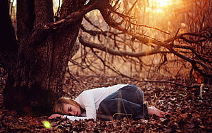 depth of field photography of woman in white long-sleeved shirt and blue jeans lying down on ground near leafless tree during golden hour