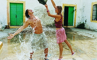 woman in red crop top and pink bottoms pouring water on man in gray shorts near white concrete house at daytime