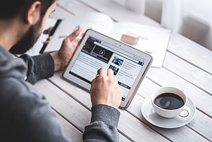 photo of man wearing gray jacket while holding turned on 10.1 Samsung Galaxy Tab 3 displaying paused video near white ceramic cup on white saucer