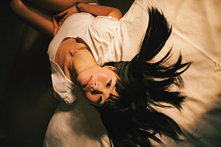 black haired lady wearing white scoop neck shirt sitting on white bed