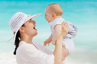 woman in white sun hat  and dress holding a baby near body of waters during daytime HD wallpaper