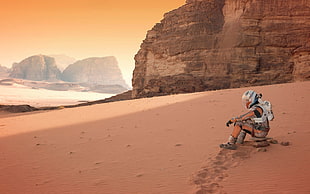 man in orange suit sitting on sand near rock