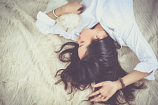 woman in white dress shirt lying on white bed spread beside white puppy
