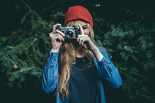 woman wearing blue denim jacket holding gray film camera standing under pine tree background HD wallpaper