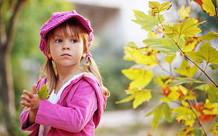 girl wearing pink zip-up hooded jacket and pink cap stands beside a green leaf plant