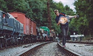 selective focus photo of man walking on railroad while holding guitar during daytime