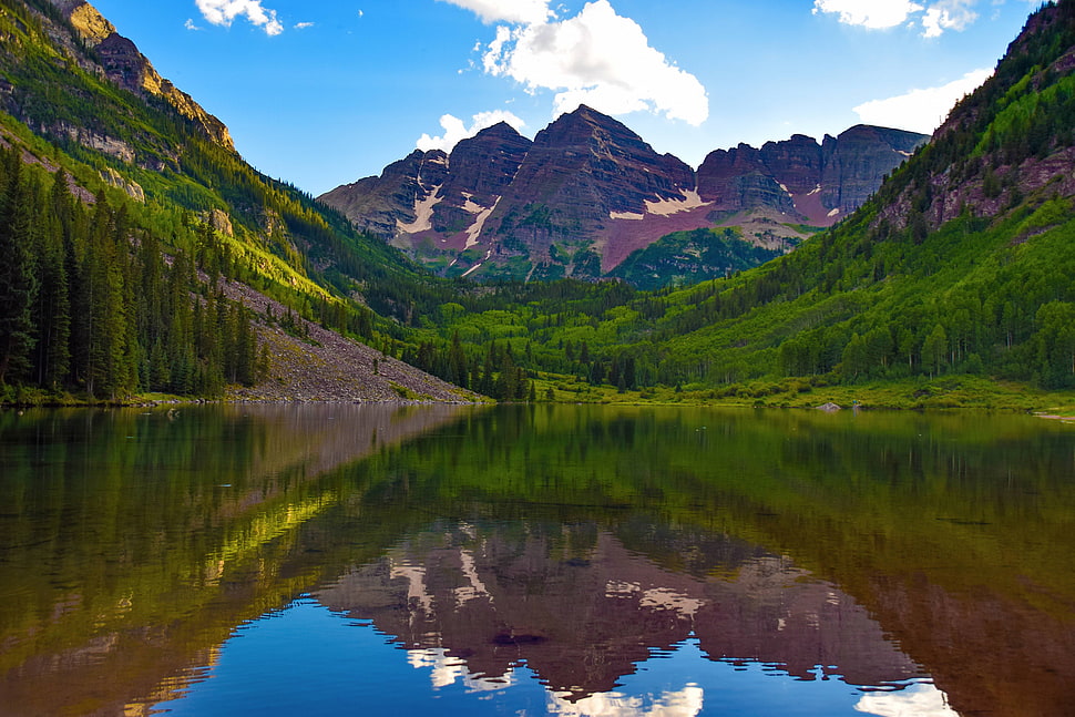 Lake Between Green Trees Across Brown Mountains Under White And Blue