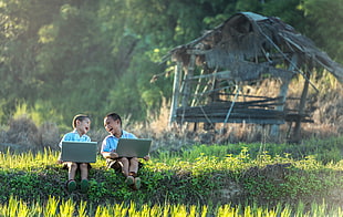two boy using laptops in front of a brown nipa hut