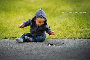 baby in black zip-up hoodie sitting on gray concrete pavement near green grass at daytime