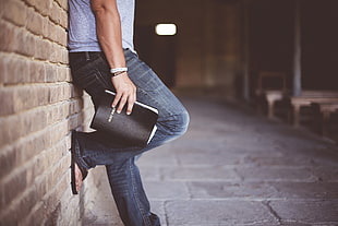 man in blue denim jeans and grey shirt holding black book leaning on brick wall