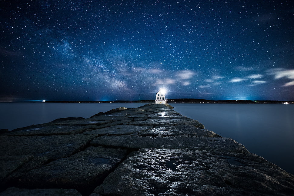 lighted white lighthouse on the edge of rock pavement under blue sky at nighttime, landscape HD wallpaper