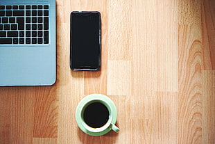 green ceramic mug on saucer beside black android smartphone and laptop computer