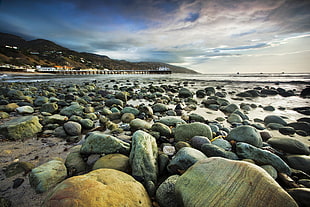 landscape photo of beach with many rocks near the dock during daytime under cloudy sky