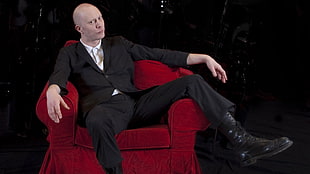 man in black formal suit sitting on red armchair
