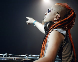 photo of man with orange braided hair wearing black framed sunglasses and black headphones pointing