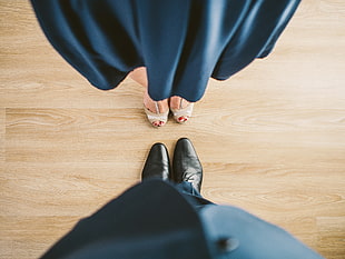 woman wearing black satin pleated dress with pair of peep-toe pumps in front of man wearing black suit with pair of dress shoes