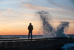 silhouette of a man standing beside body of water with water splash during sunset