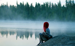 photo of a red haired female sitting on rock near body of water