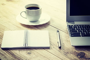 silver and black laptop computer, white ruled paper, pen, ceramic coffee cup and saucer on top of beige wooden surface
