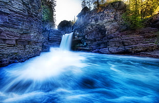 blue water running through falls on gray rock formations  with green leaved trees HD wallpaper