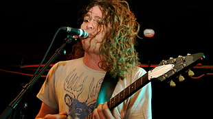 man wearing gray t-shirt holding electric guitar close to microphone with stand