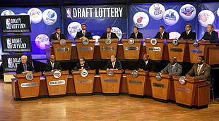 men wearing suits sitting on brown wooden desk on stage with Draft Lottery on background