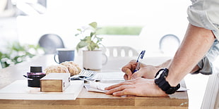 person holding pen leaning on wooden table with paper and mugs