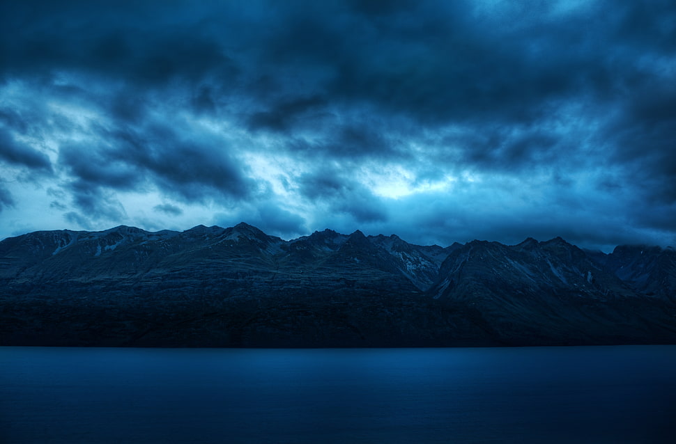 rocky mountain beside body of water under blue and black sky HD wallpaper