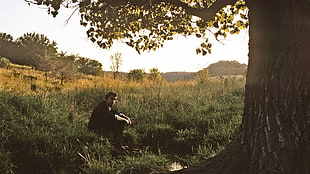 man wearing black shirt and black pants sitting next to tree and grass during daytime