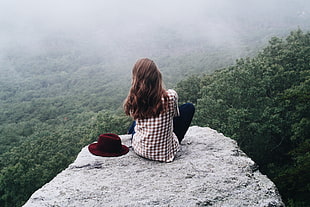 woman wearing check print top sitting on the edge of the gray boulder rock beside the red hat overlooking the green forest HD wallpaper